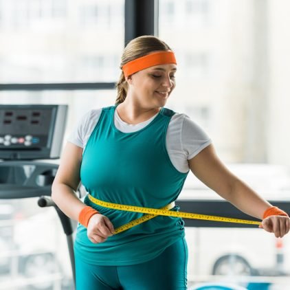 cheerful plus size girl looking at measuring tape while measuring waist in gym