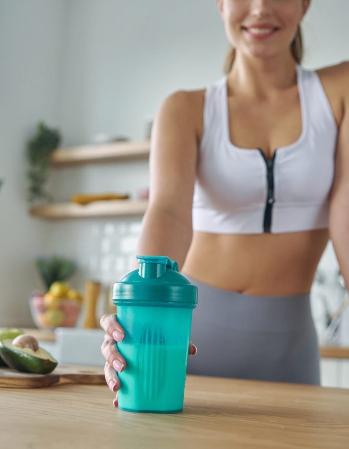 Close-up of woman in sports clothing holding shaker with protein cocktail at the kitchen